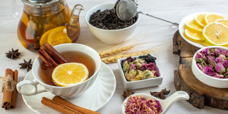 A white tabletop display featuring a cup of tea with a lemon slice and cinnamon stick, dried rosebuds, star anise, and a glass teapot.