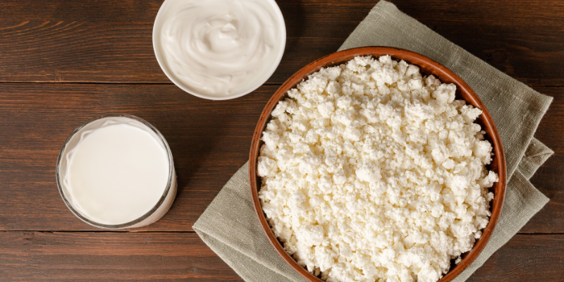 A wooden table featuring a large bowl of fresh curd cheese, a glass of milk, and a small bowl of creamy kefir or yogurt.