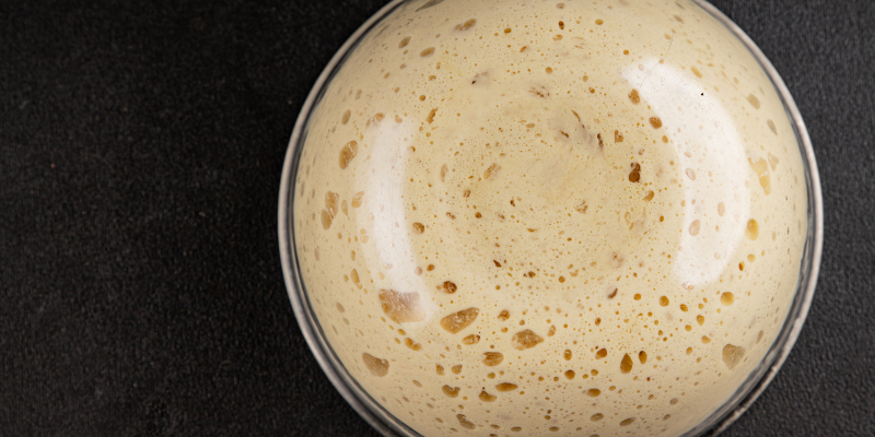 A top-down close-up of a bubbly, frothy yeast starter in a glass bowl, showing active fermentation with many small air pockets.
