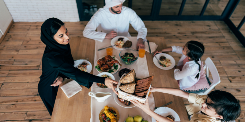 A family sits around a long wooden table sharing a meal of roasted chicken, salads, and bread, with glasses of orange juice and tea.
