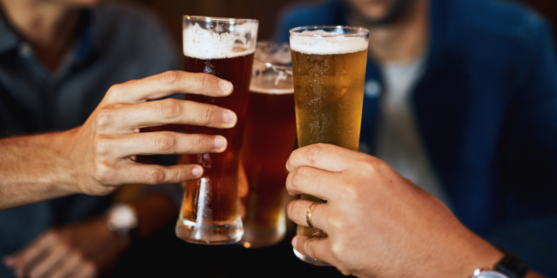 A close-up of three people clinking glasses of various types of beer in a dimly lit social setting, with two individuals smiling in the soft-focus background.