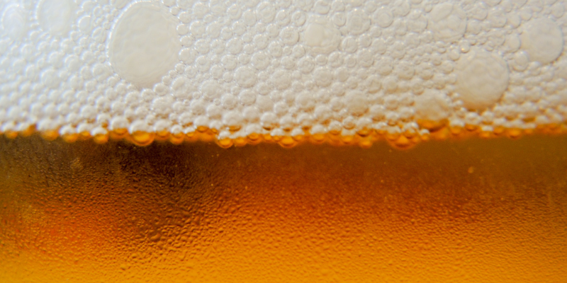 A macro photograph showing the fine, white bubbles of beer foam against a golden, carbonated liquid background.