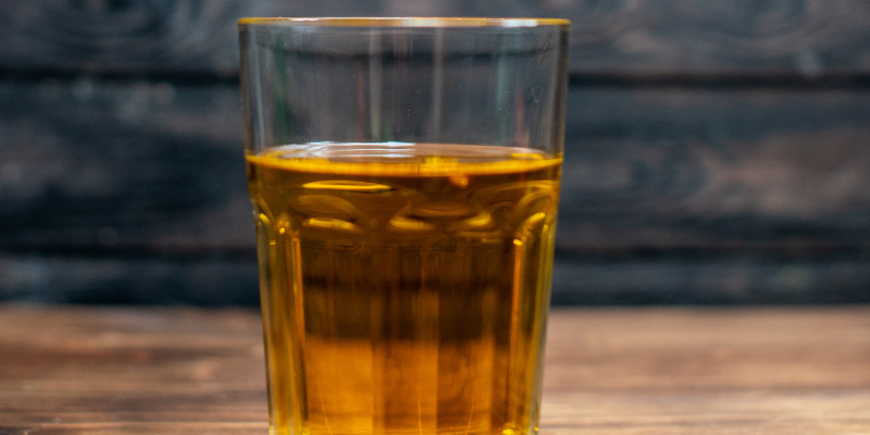 A hand dipping a thin round apple slice into a glass of golden apple cider on a dark wooden background.