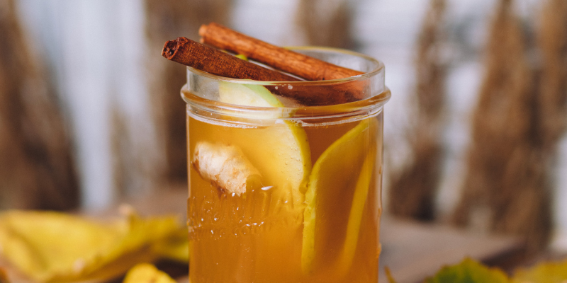 A glass jar filled with amber Tepache drink, garnished with a cinnamon stick and fruit slices on a wooden table.