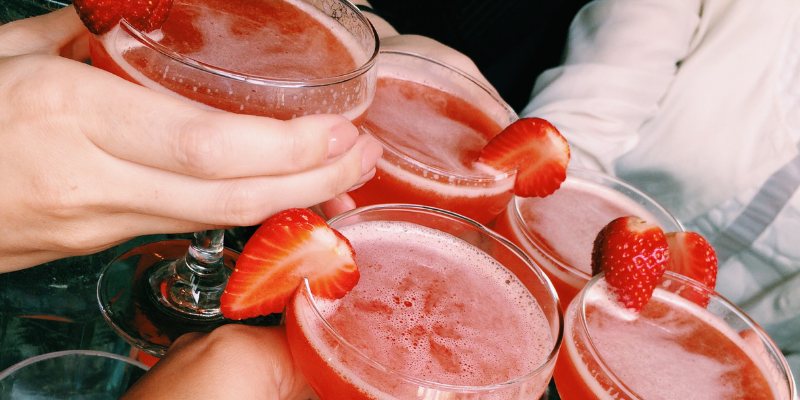 A group of friends toasting with glasses of pink strawberry shrub drinks garnished with fresh berry slices.
