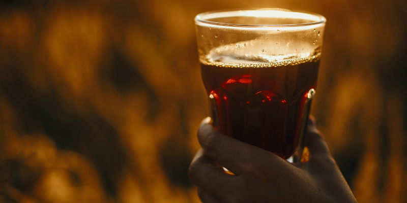 A hand holding a glass of dark, amber-colored sarsaparilla drink against a golden, sunlit background.
