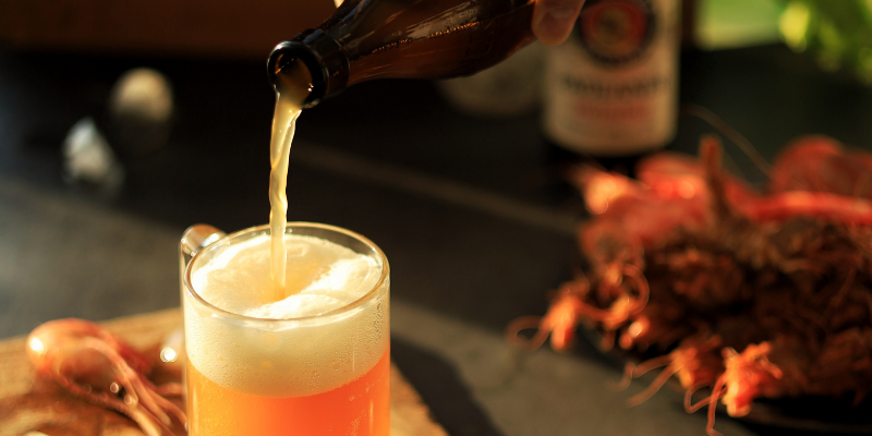 A hand pouring a golden, carbonated beer from a bottle into a glass mug on a dark tabletop.