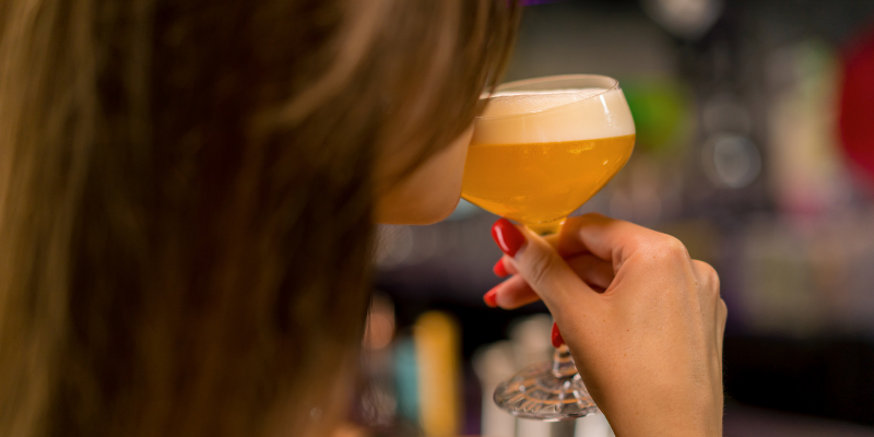 A close-up view from behind a person holding a stemmed glass of golden beer to their lips in a dimly lit bar setting.