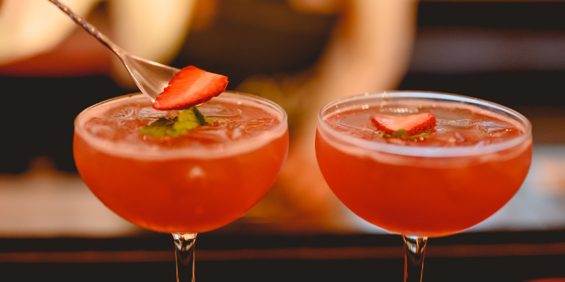A bartender garnishing two bright red cocktails with fresh strawberry slices using a cocktail spoon.