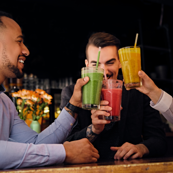 A cheerful group of friends sitting at a bar table, clinking glasses filled with green, red, and yellow non-alcoholic smoothies and juices.