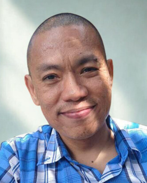 A portrait of Bryan Samoy, the alcohol-free brewing expert and writer for AlcoFreeBrew, standing in a dimly lit traditional pub. He is a bald man with a grey beard, wearing a light green button-up shirt and looking directly at the camera.