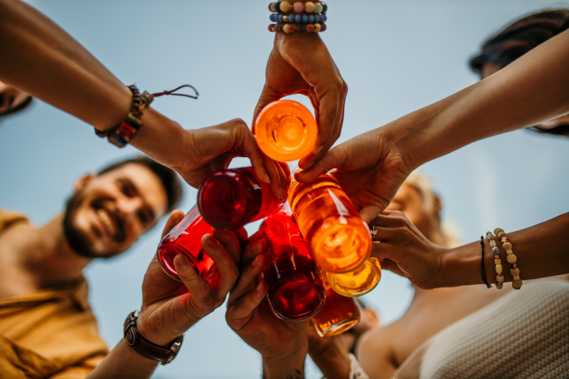 A low-angle view looking up at a circle of friends' hands toasting with colorful red and orange glass bottles against a clear blue sky.