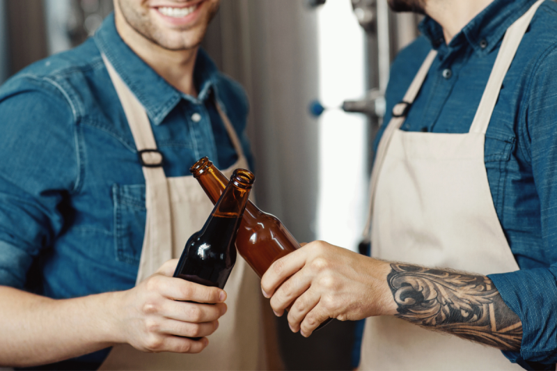 Two brewers in denim shirts and aprons clinking brown beer bottles together in a celebratory toast.