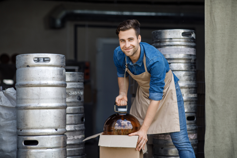 Jonas Leroy crouching down to unpack a large glass carboy (demijohn) fermentation vessel from a cardboard box, looking ready to start a new batch.