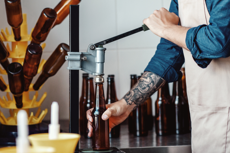 Close-up of a brewer's hands using a manual lever capper tool to seal a brown glass beer bottle on a workbench.